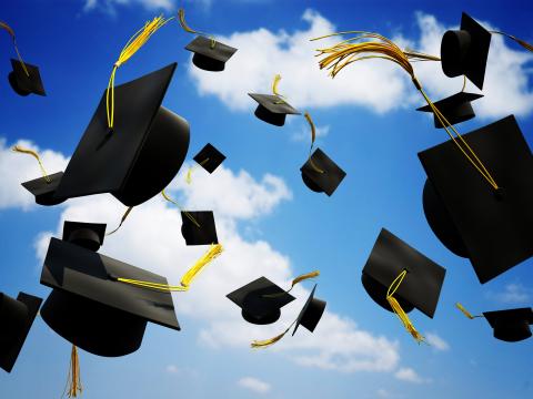 Graduation caps with tassels are thrown in the air set against a partly cloudy sky.