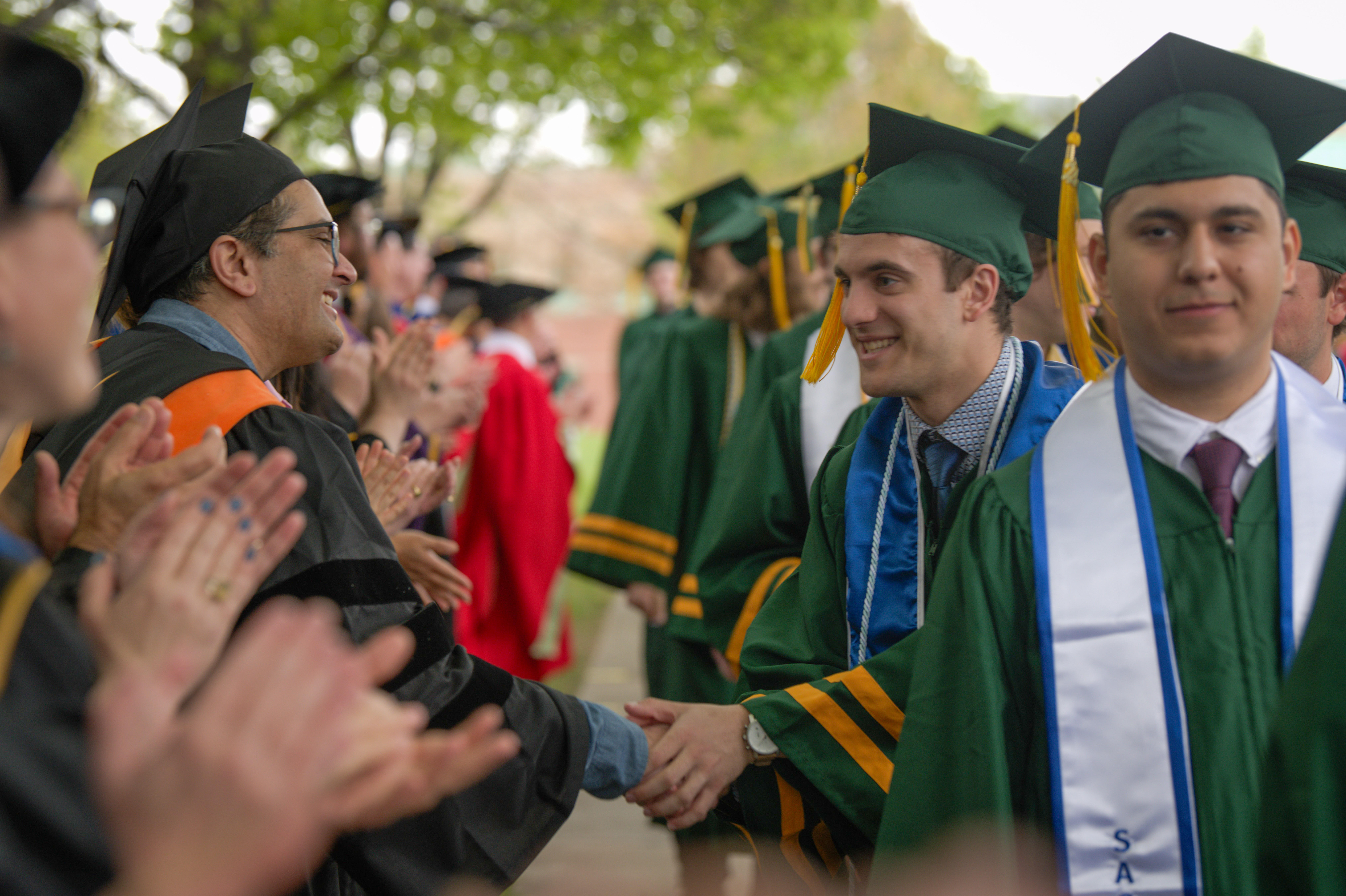 A student in regalia processing with peers shakes hands with a faculty member in regalia on as the students passes by applauding faculty