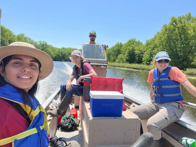 Four biology student researchers ride in a boat on a river with blue sky in background