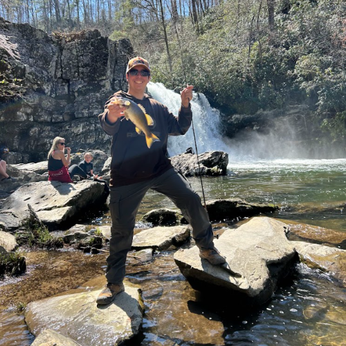 A photo of Robert Greenberg holding a fish in front of a waterfall