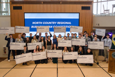 Wide group photo of 32 North Country Regional Business Plan Competition winners holding over-sized checks in the air with a large display in background reading "North Country Regional Business Plan Competition"