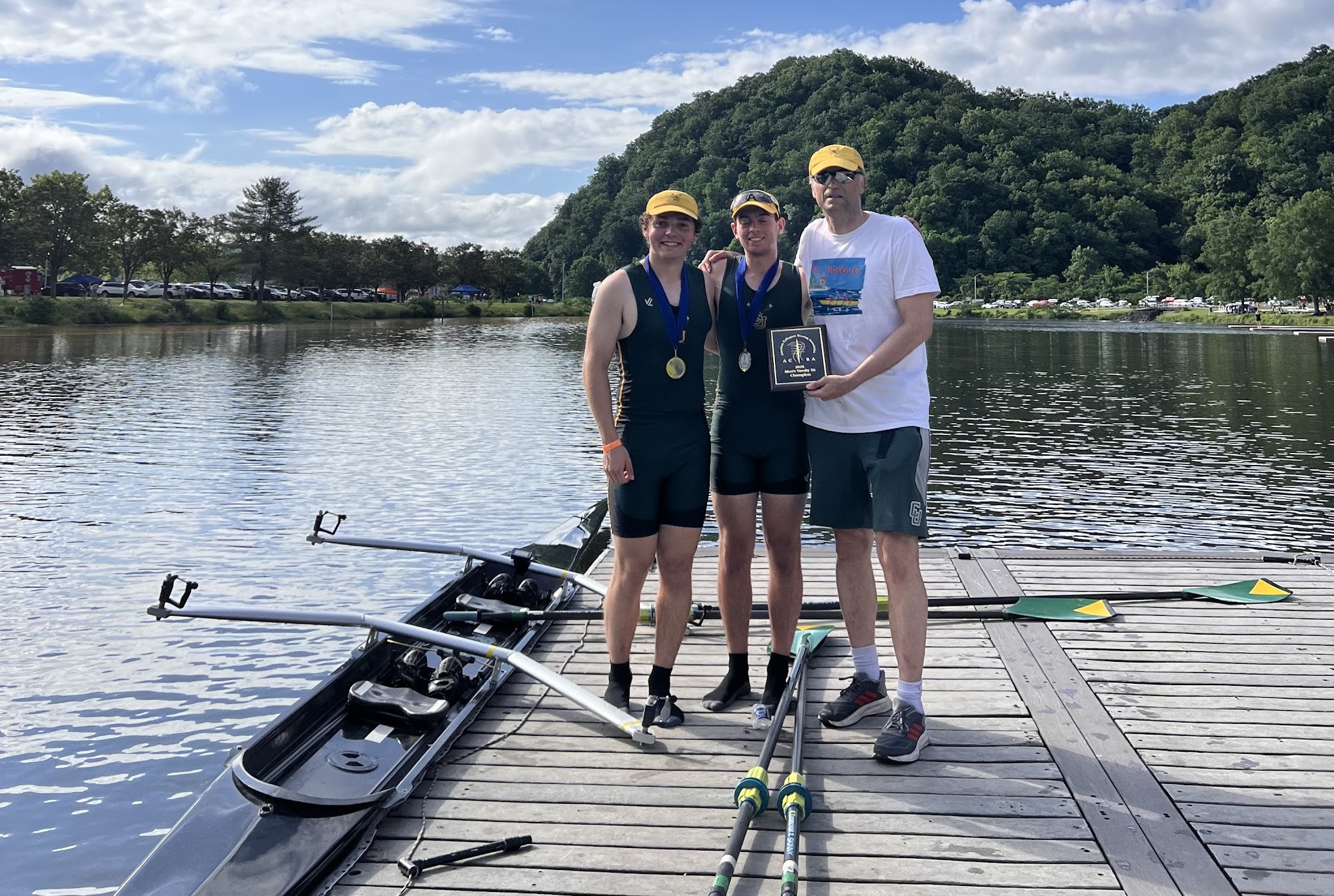 two crew athletes pose for a photo with their coach on a dock with their boat in the water next to them