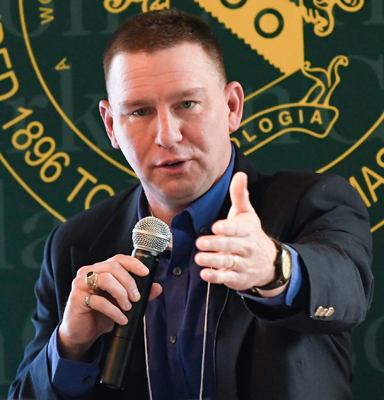 Erik Backus in black suit jacket and dark blue open-collared shirt holding a microphone in his right hand and extending his left hand forward, with part of a green and gold Clarkson University seal in the background.