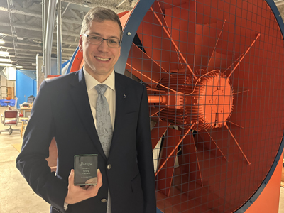 Portrait of Craig Merrett holding a crystal plaque next to a large fan connected to a wind tunnel