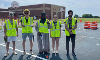 Five students pose for a photo in high-visibility vests on an outdoor basketball court with various traffic cones in the background