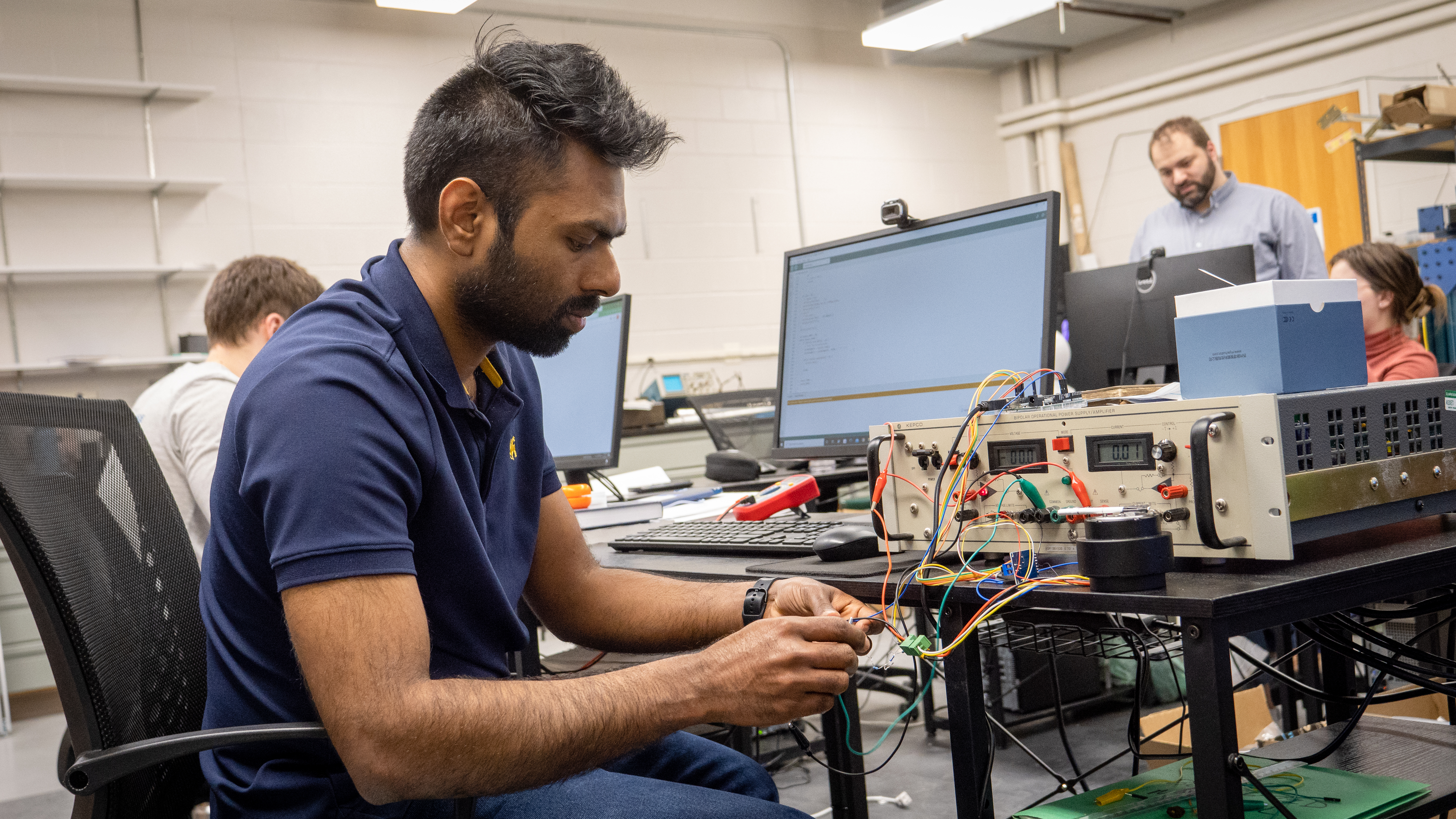 A student sits at a computer connecting wires to a computer hardware box in a lab. 