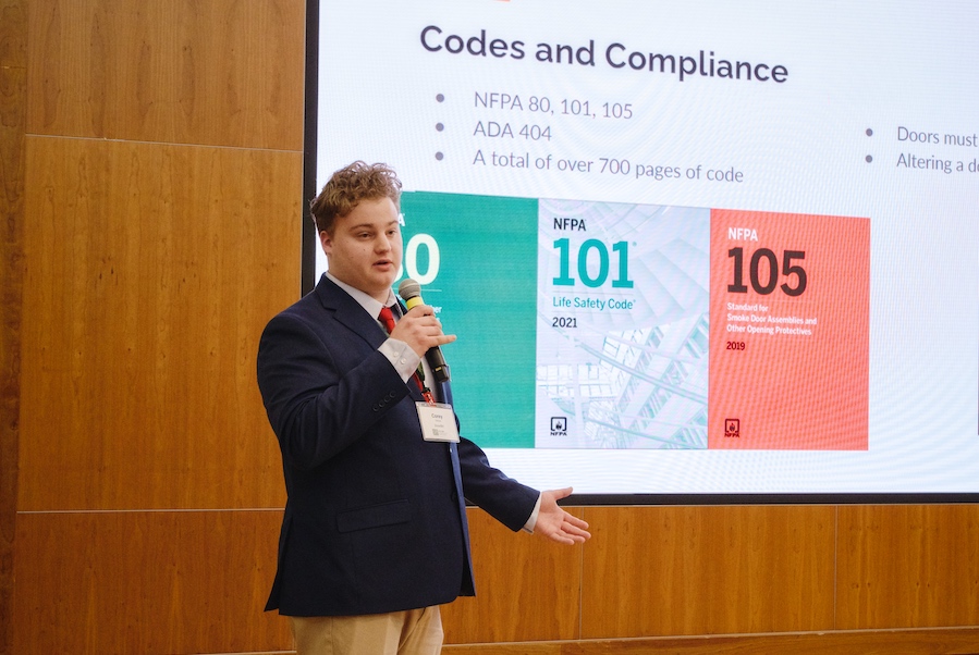 A male student, wearing business attire, speaks into a microphone while standing in front of a large screen.