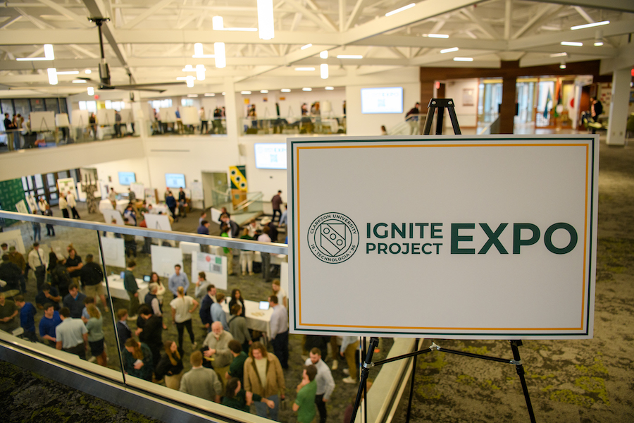 At Clarkson University a sign reading “Ignite Project Expo” is displayed on a balcony, while students, faculty, and staff can be seen interacting near poster boards on the floor below.