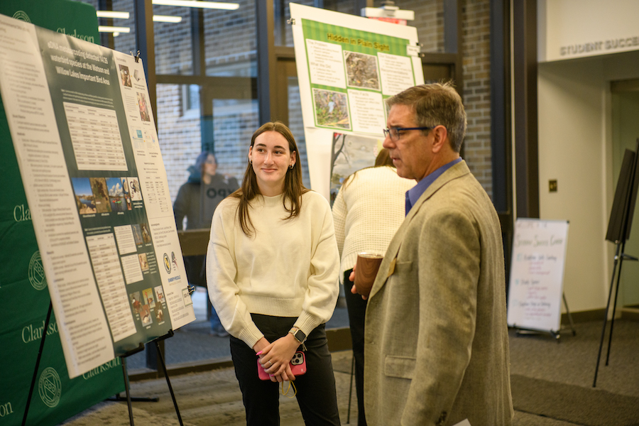 A female student smiles while a male professor reads her poster board.