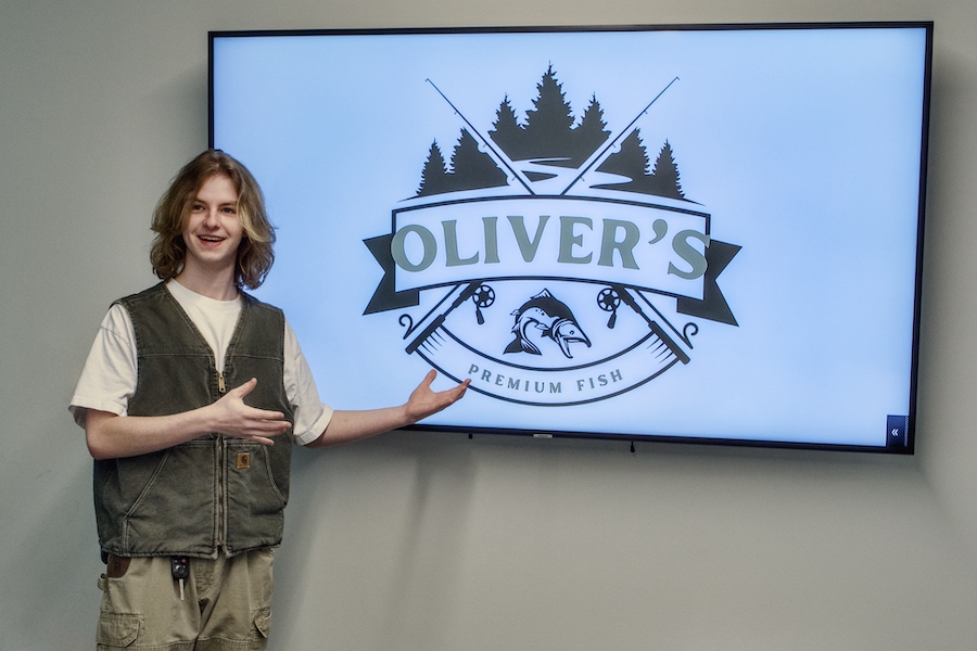 A male student smiles while pitching for a business, with the words “Oliver’s Premium Fish” shown on an electronic screen.