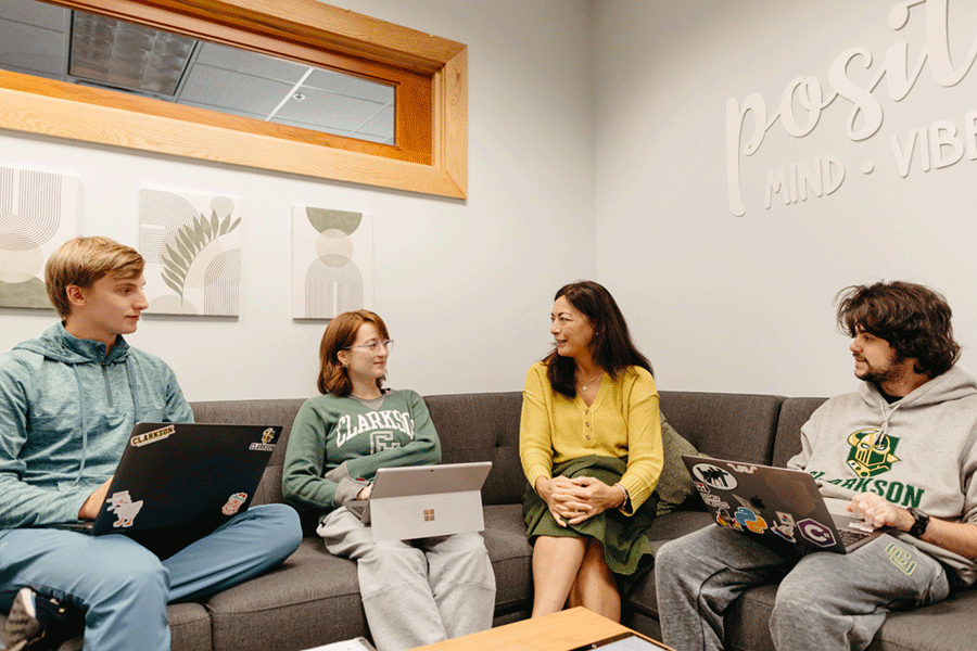 A Clarkson staff member (second from right) sits on a couch and chats with three Clarkson students who have their laptops on their laps.