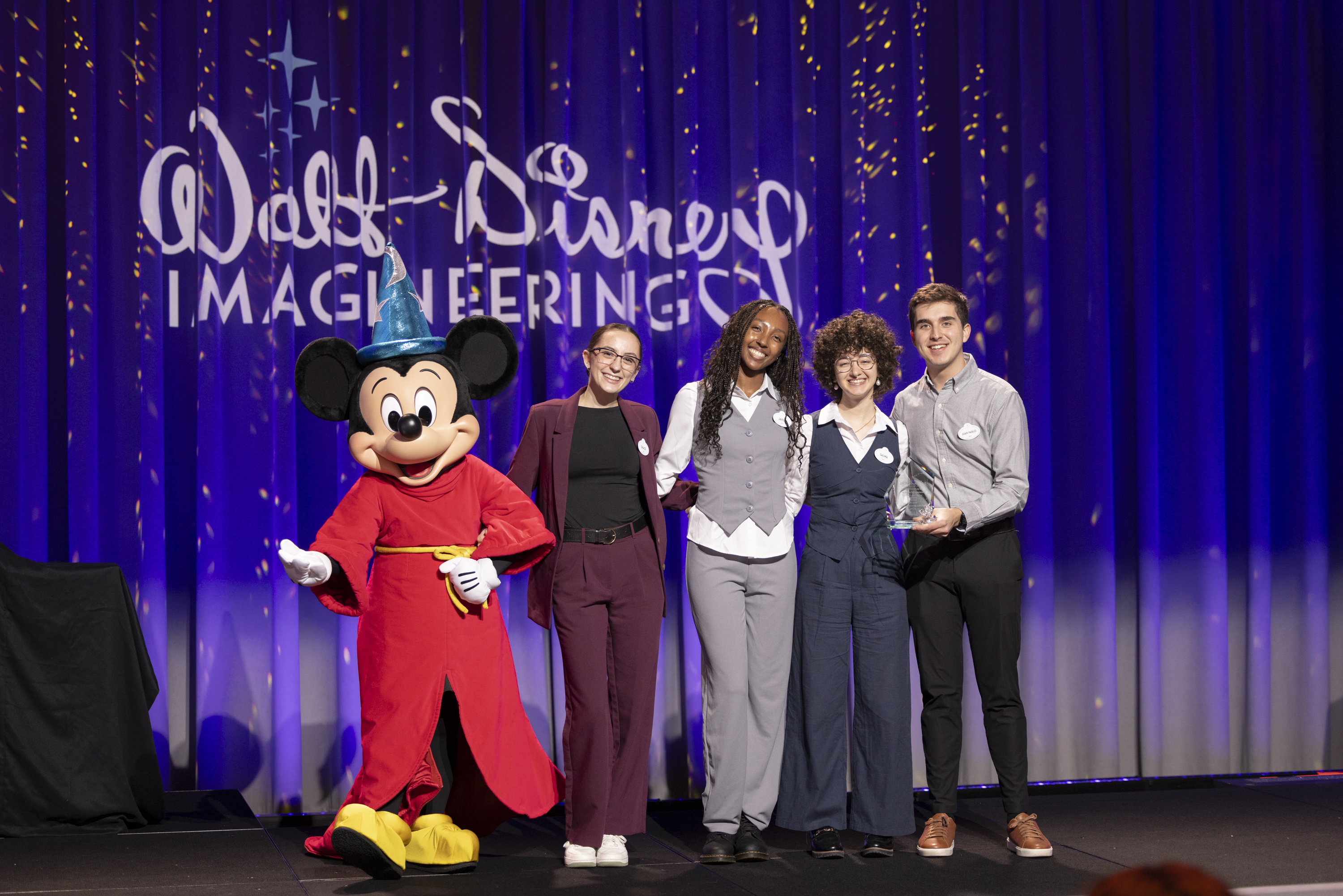 Four students stand on a stage with Mickey Mouse, holding an award for winning a competition. 