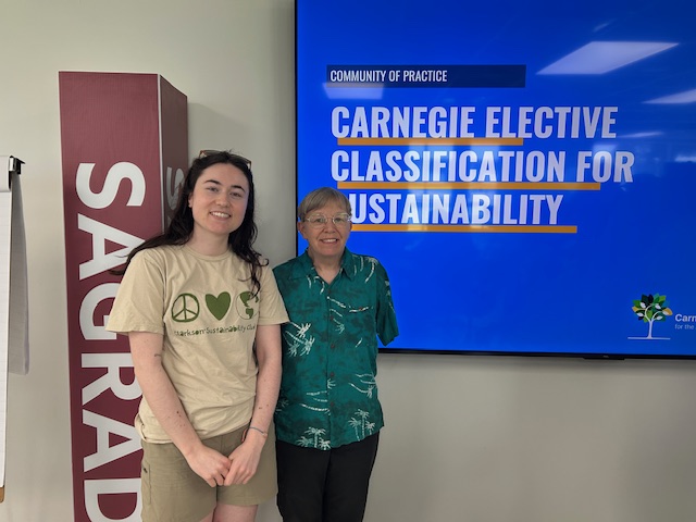 Two women pose for a photo next to a television screen that reads "Carnegie Elective classification for sustainability"