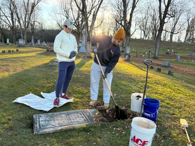 Two students stand next to a downed headstone while one uses a shovel to dig a space for the headstone