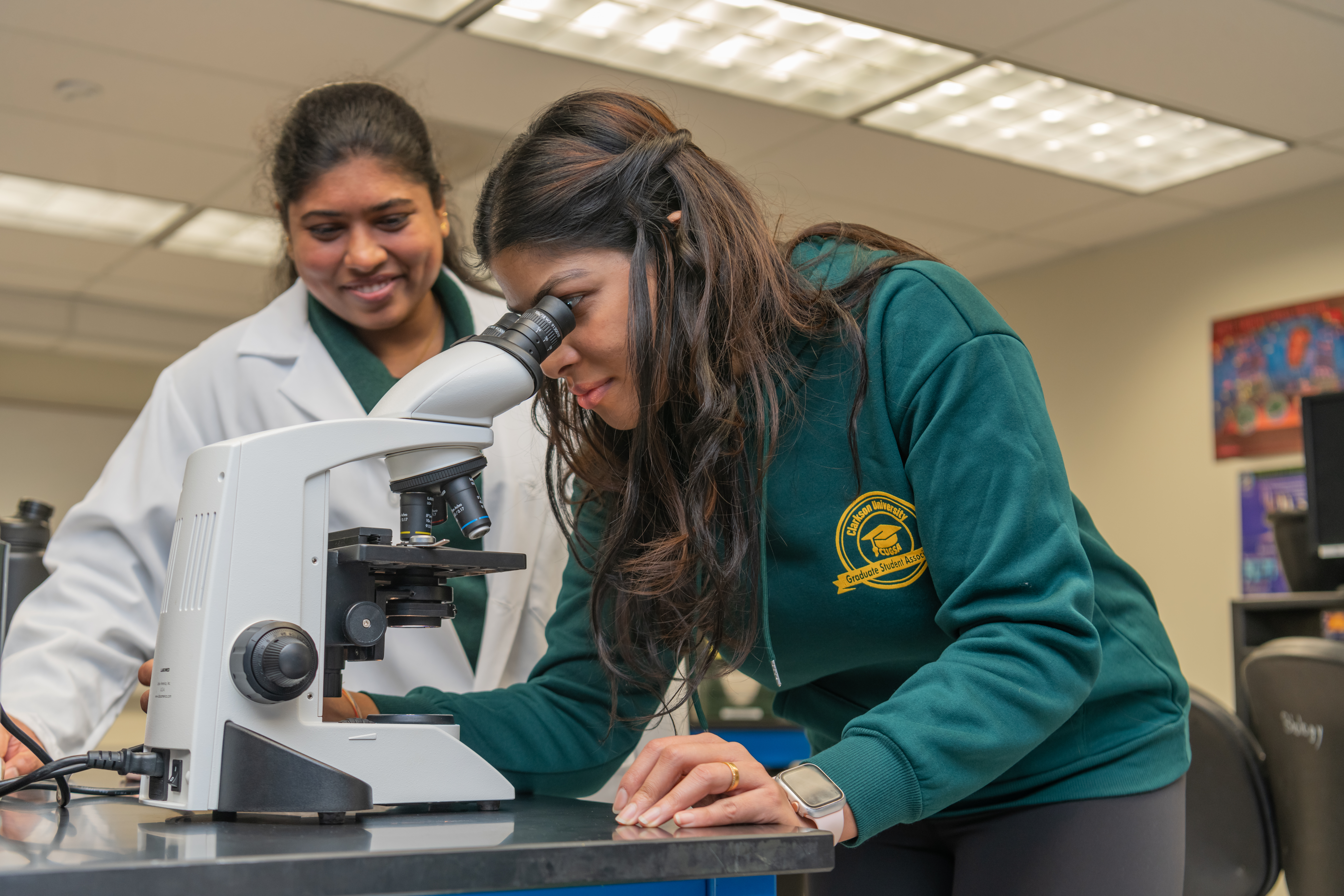A student leans down to look through a microscope while a second student in a lab coat supervises nearby