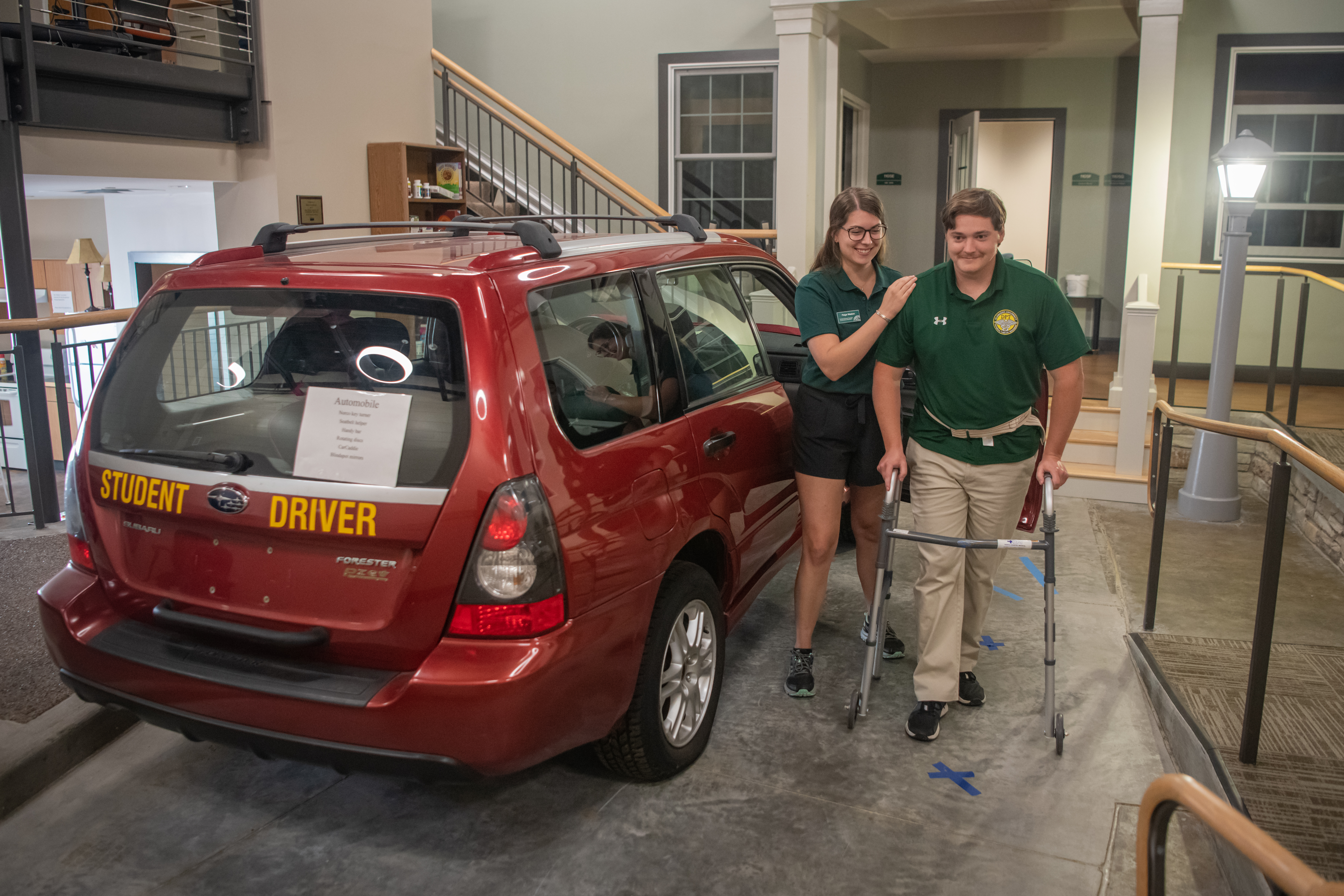 A physical therapy student simulates a patient using a walker next to a vehicle in a simulation lab as another student practices aiding the patient
