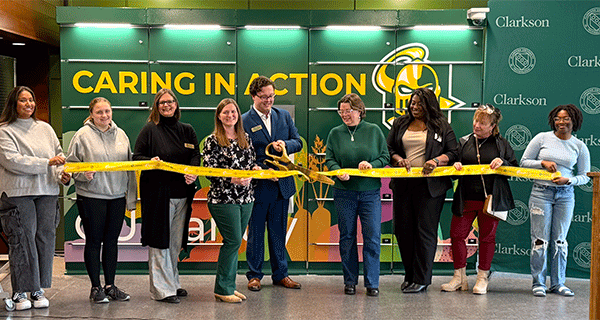 Clarkson students and staff of the CU Pantry hold a gold ribbon and stand in front of food lockers that reads "Caring in Action" with the Golden Knight logo led by leader Johannes Richter cutting a gold ribbon