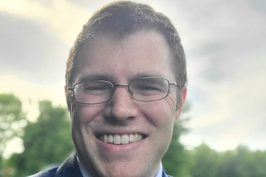 A professional headshot of Professor Ian McCrum, who is smiling and wearing glasses and a blue suit jacket. He is pictured outdoors with a soft-focus background of green trees and a bright sky.