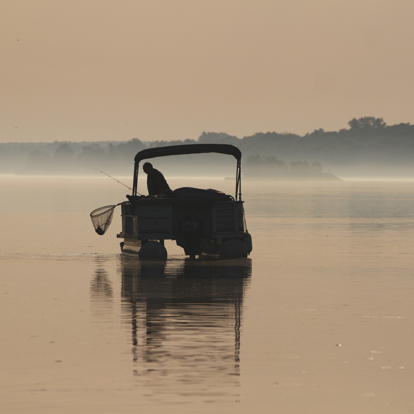 A silhouette of a person fishing from a pontoon boat on the calm, misty St. Lawrence River at dawn. The water is perfectly still, reflecting the boat and the soft, hazy sky in shades of sepia and gold.