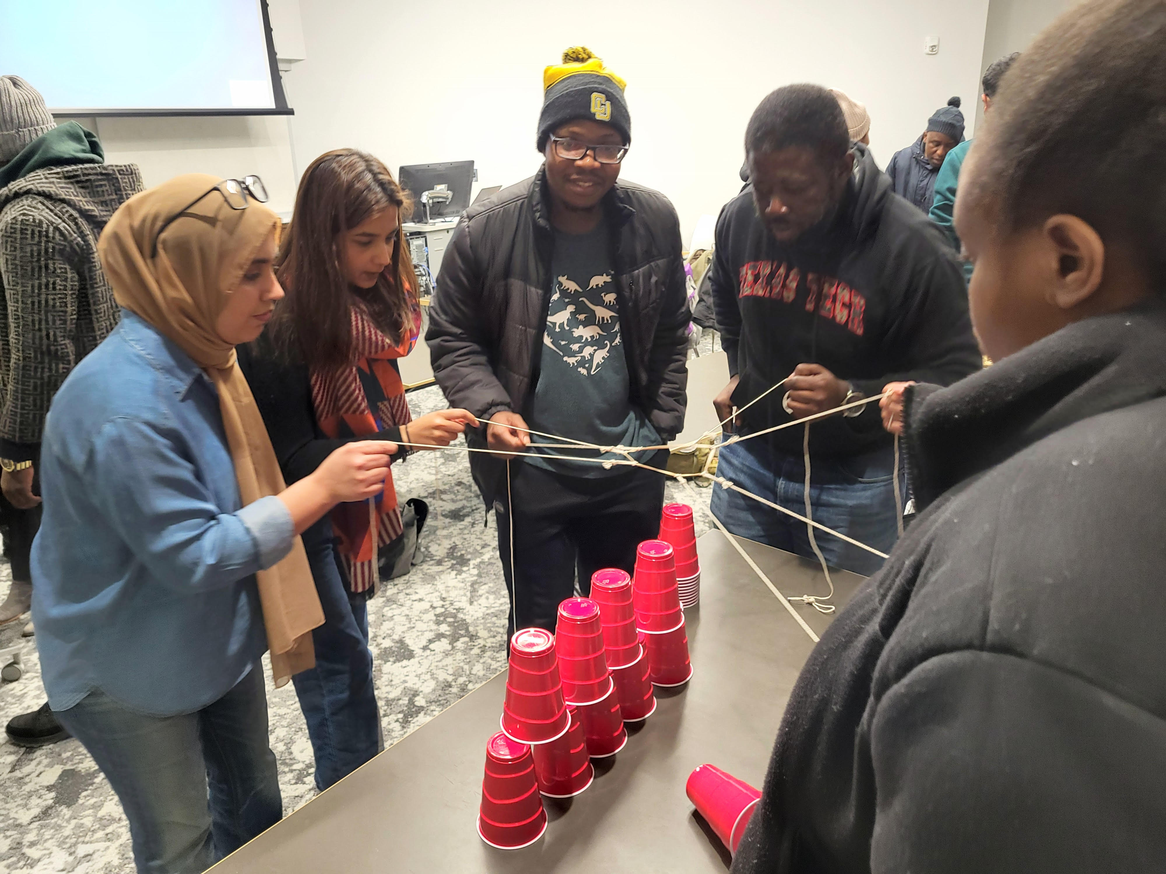 Students stand in a circle holding strings that are all tied to a loop, which they use to try and collectively pick up a cup.