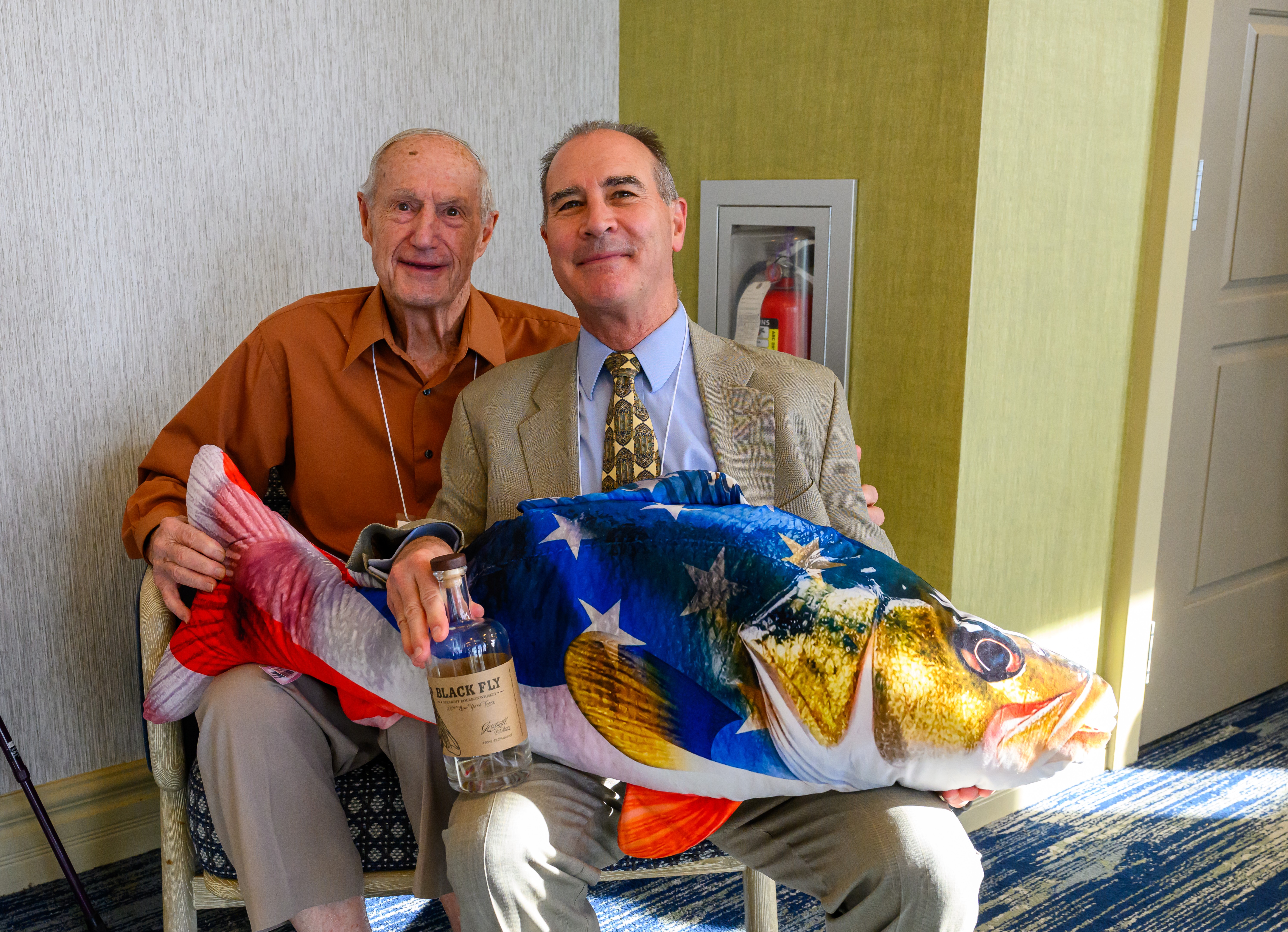 Two people sit together at an event, smiling toward the camera while holding a large decorative fish-shaped pillow featuring an American flag design. One person also holds a bottle labeled “Black Fly.” The scene appears to be a social gathering or conference reception, with a small table, refreshments, and a fire extinguisher cabinet visible in the background. Sunlight streams into the room, illuminating the subjects and the colorful fish decoration.