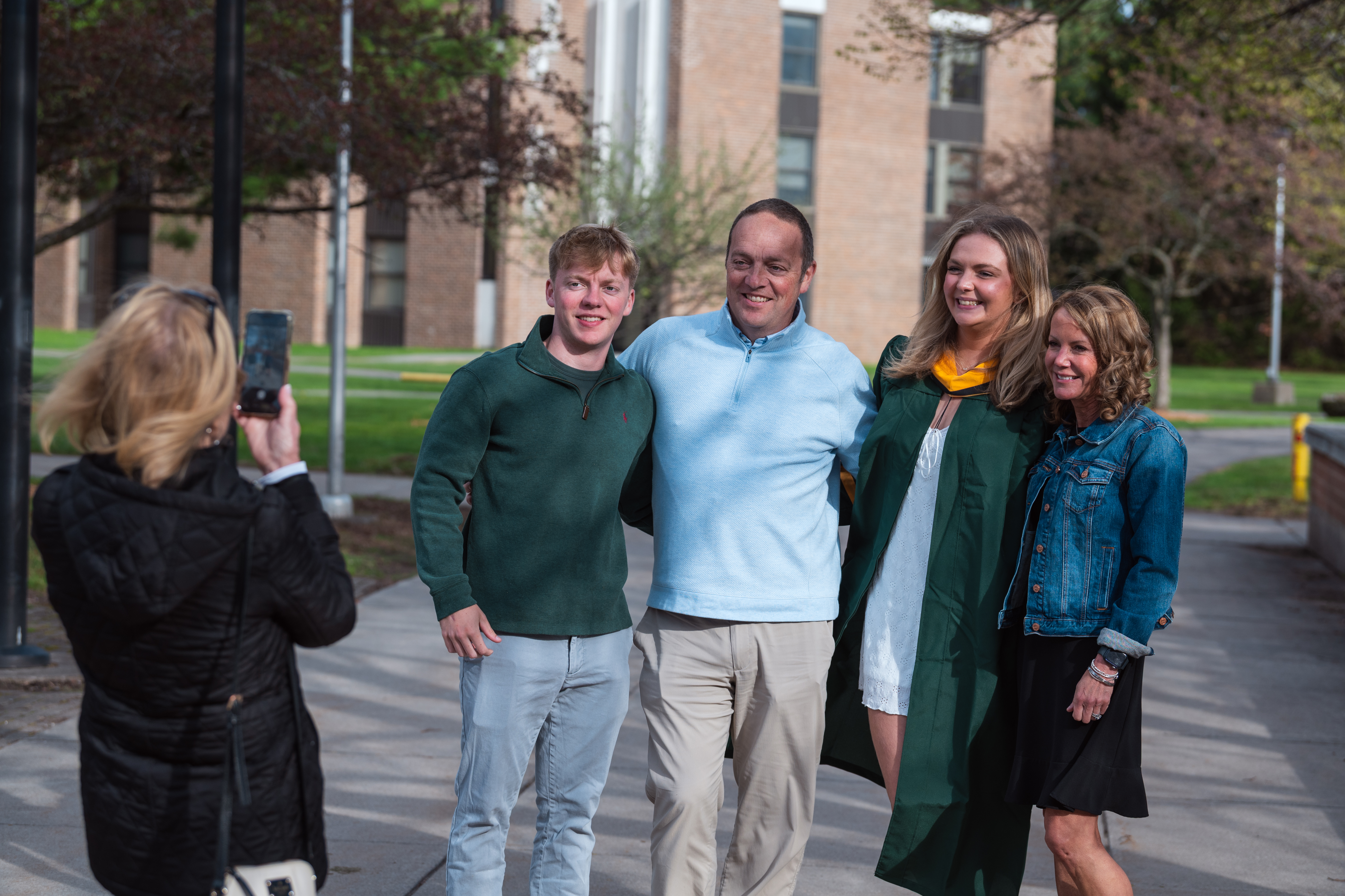 A group of four people pose outdoors for a photo on a college campus while another person takes their picture with a smartphone. One person in the group is wearing a green graduation gown with a gold hood, while the others are dressed in casual clothing. They stand together smiling on a walkway with campus buildings, trees, and lawns visible in the background.
