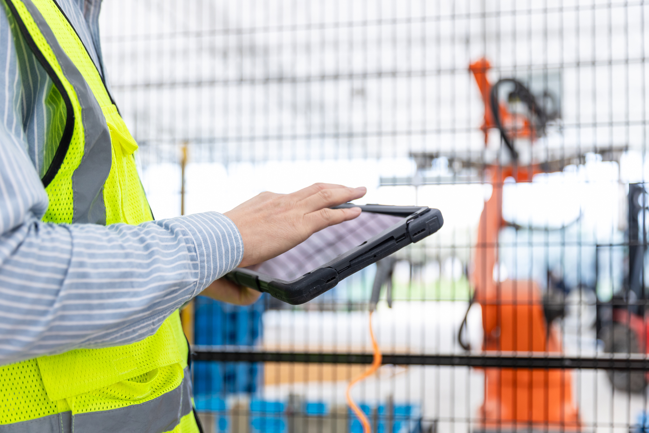 Close-up of a person wearing a high-visibility safety vest using a rugged tablet device in an industrial setting. A wire safety fence and blurred manufacturing or automation equipment are visible in the background, suggesting monitoring, inspection, or data collection activities.