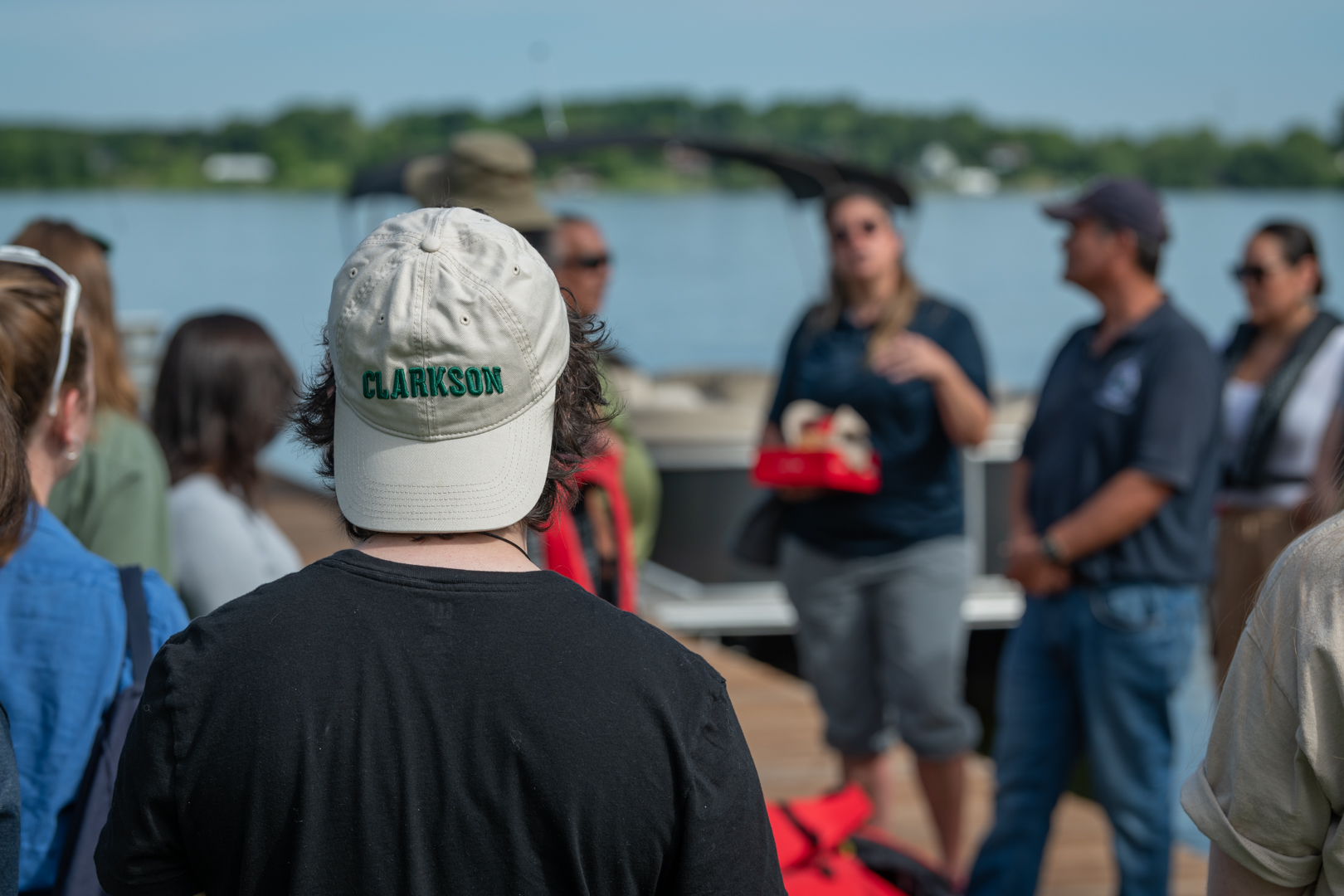 A group gathers on a dock beside a lake for an outdoor research activity. In the foreground, a participant wears a cap embroidered with “Clarkson” while listening to presenters near a boat.