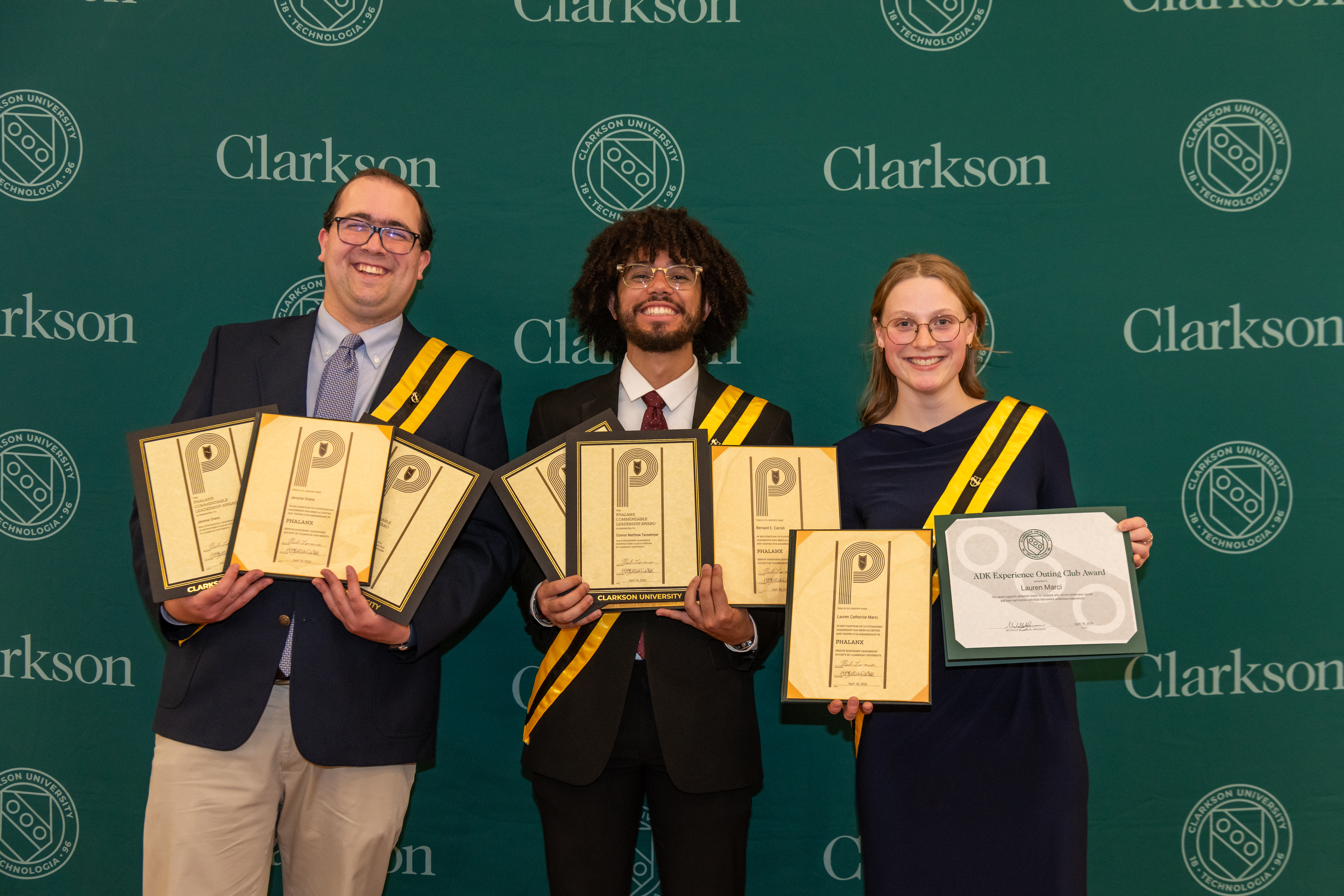 Three smiling Clarkson University students wearing academic sashes stand in front of a green backdrop holding multiple framed award certificates.