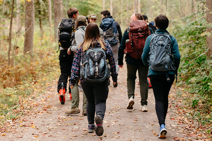A group of Clarkson students wearing backpacks are walking on a wooded trail